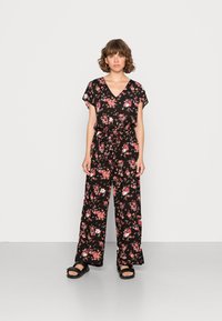 Jumpsuit negro con flores, mangas cortas, escote en V, cinturilla con lazo y pantalones de pierna ancha, con patrones de flores rosas y blancas. Sandalias negras.