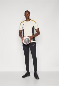 Conjunto deportivo de Nike que presenta una camiseta de manga corta color crema con acentos en negro y dorado, combinada con pantalones negros ajustados y zapatos negros.