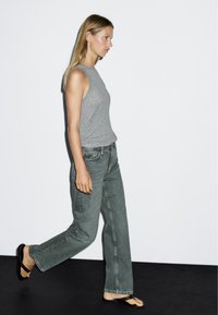 Gray sleeveless top, faded green wide-leg jeans, and brown flip-flops. Model walking against a plain white background.