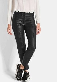 Schwarze Lederhosen mit einem taillierten Design, vorderen Nähten und knöchellanger Länge, kombiniert mit schwarzen Sneakers, vor einem neutralen Hintergrund.