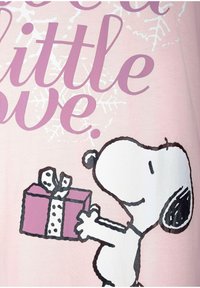 Light pink t-shirt featuring Snoopy holding a purple gift box with a white ribbon. Text reads "Need a little love" in purple.