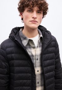 Νέος άνδρας με σγουρά μαλλιά φορώντας μαύρο φουσκωτό μπουφάν πάνω από πουκάμισο σε μπεζ και μαύρο καρό, με φόντο ένα απλό λευκό τοίχο.