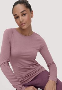 Young woman with tied-back hair wearing a long-sleeve mauve top and purple pants, sitting and looking softly at the camera.