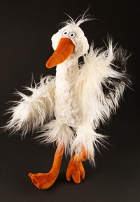Plüschreiher-Spielzeug mit einem flauschigen weißen Körper, orangefarbenem Schnabel und Beinen. Schwarze Akzente an den Flügeln und eine Haarsträhne oben. Große cartoonartige Augen.