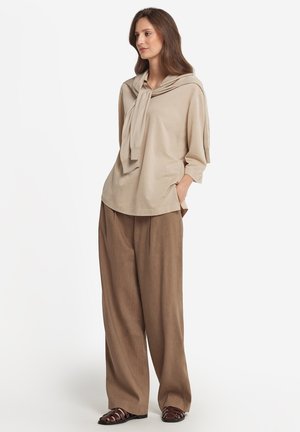 Beige lange mouwen top met een geknoopte halslijn, gecombineerd met losse, bruine wijde broekspijpen en bruine sandaaltjes met banden. Zachte texturen overal.