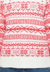 Suéter branco com padrões nórdicos vermelhos, incluindo flocos de neve e formas geométricas, usado por dentro de jeans de ganga azuis.