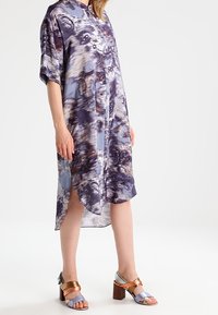 Hemdkleid aus leichtem Stoff mit einem blau-lila abstrakten Muster, ausgestattet mit einer Knopfleiste und kurzen Ärmeln. Kombiniert mit farblich abgesetzten Absätzen.