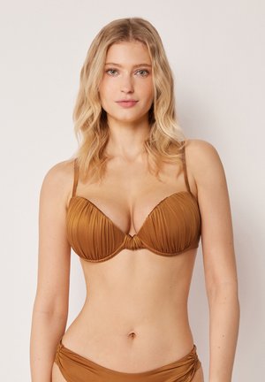Blonde Frau mit welligem Haar, die ein braunes, gerafftes Bikinioberteil und passende tiefe Bikinihosen vor einem schlichten Hintergrund trägt.