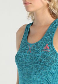 Una camiseta sin mangas turquesa con un patrón texturizado, que presenta un logo de acento rojo y un diseño en forma de X en el frente, hecha de un material elástico y transpirable.