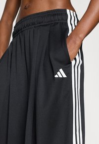 Μαύρο αθλητικό παντελόνι με πιετέ σχέδιο, που διαθέτει λευκές ρίγες κατά μήκος των πλευρών και λογότυπο Adidas στ�ην εμπρόσθια τσέπη.