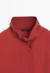 Red button-up shirt featuring a structured collar, double-button placket, and smooth cotton fabric with a slight sheen. Brand label visible.