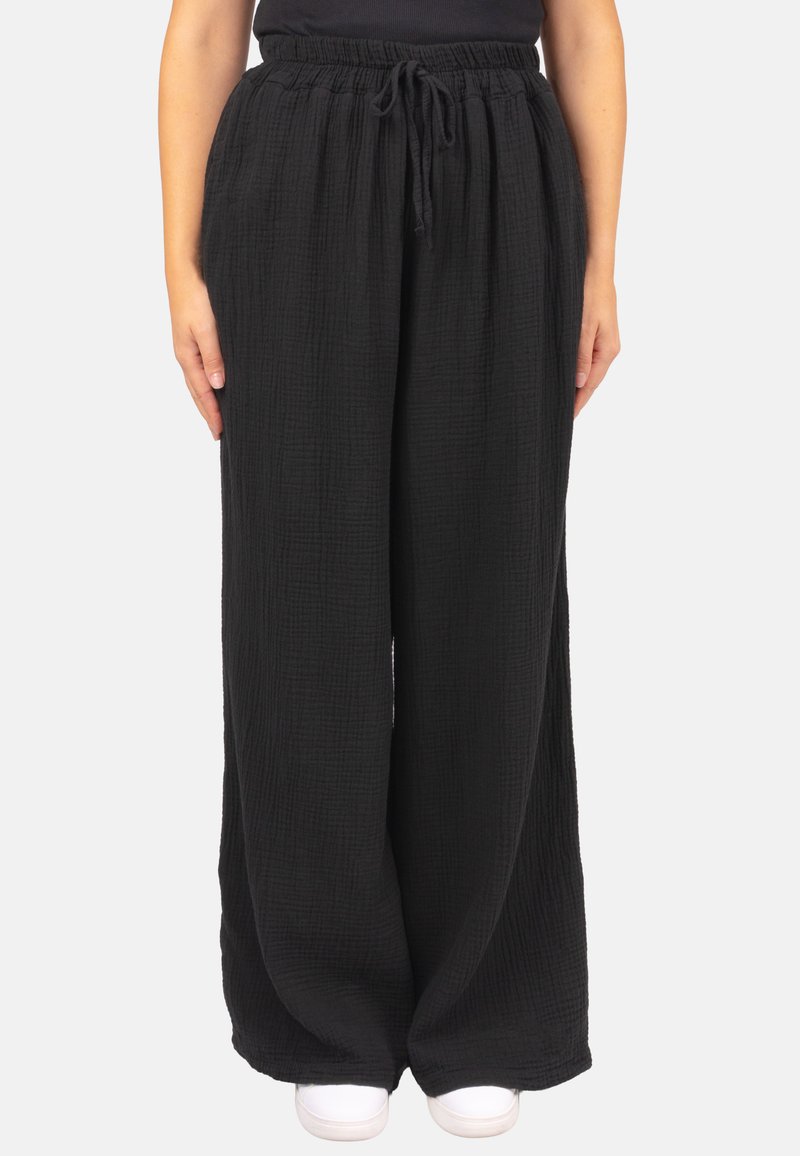 Schwarze, weite Hose aus strukturiertem Stoff. Verfügt über einen elastischen Bund mit Kordelzug. Das Model trägt dazu weiße Sneaker.