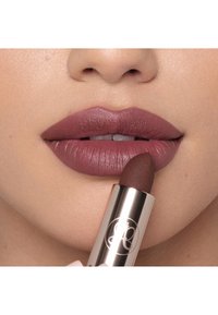 Lápiz labial mate en tono malva sostenido cerca de labios llenos; el tubo de lápiz labial es plateado metálico con un logotipo, mostrando una punta ligeramente redondeada y una textura suave.