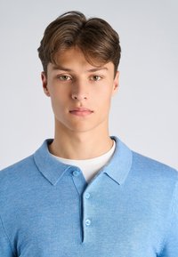 Jasnoniebieska koszulka polo o fakturowanej powierzchni, z kołnierzykiem i trzema dopasowanymi guzikami, noszona na białej koszulce z okrągłym dekoltem.