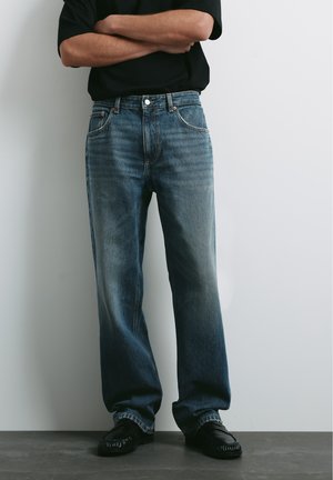Personne portant un jean bleu ample, des chaussures noires et une chemise noire, les bras croisés, debout contre un mur clair uni.