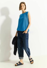 Top senza maniche blu, jeans in denim scuro con risvolti, e sandali blu navy. La modella tiene un maglione scuro, in piedi contro uno sfondo chiaro.
