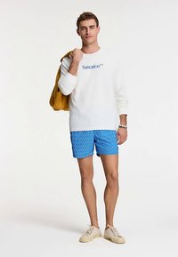 Fehér hosszú ujjú ing "Suncation™" logóval, kék mintás fürdőgatya és bézs cipő párosításával. Sárga válltáska. Vékony szabás.