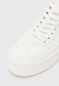 Baskets en cuir blanc avec une surface lisse, un bout rond, une semelle texturée et des lacets plats. Présente une couture tonale et un design minimaliste.