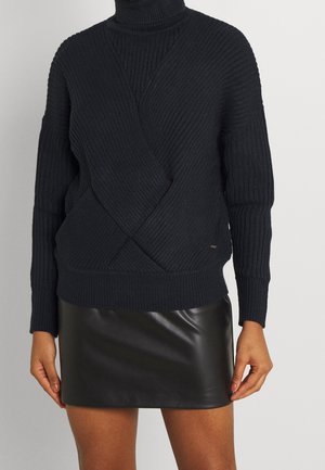 Femme portant un pull à col roulé en maille côtelée noire avec un motif croisé et une mini-jupe en cuir noire.