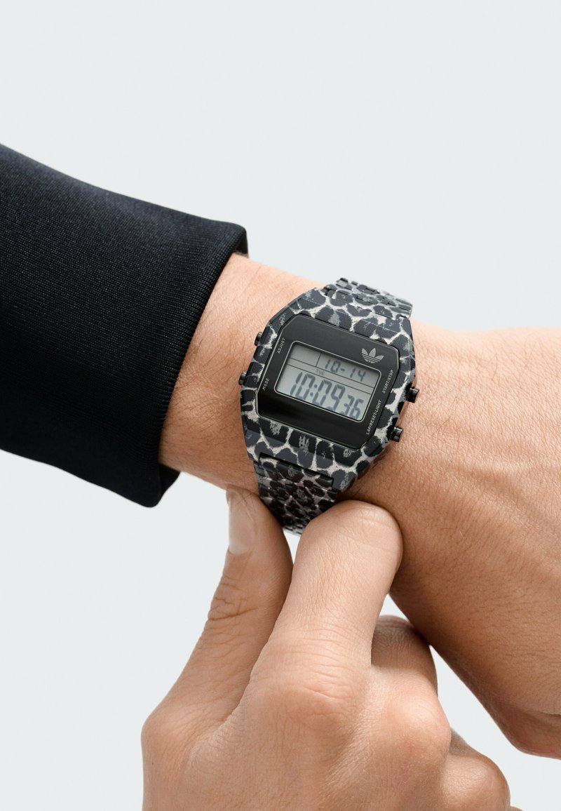 Digitale Uhr mit einem schwarzen und grauen Leopardenmuster-Armband, rechteckigem Zifferblatt und sichtbarer Zeitanzeige, getragen am Handgelenk.