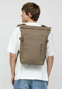 Brauner Rolltop-Rucksack mit Reißverschlusstasche, verstellbaren Trägern und einer glatten, strukturierten Oberfläche. Minimalistische Markenkennzeichnung auf der Vorderseite.