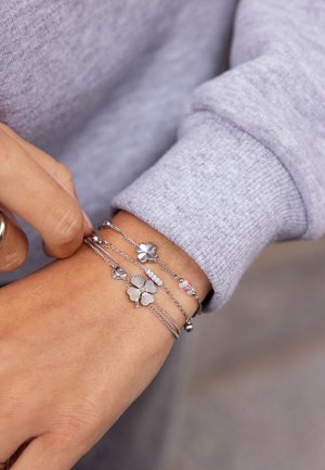 Bracciali metallici argento con accenti floreali, catene e perline in delicati colori pastello, indossati su un polso accanto a un maglione grigio.