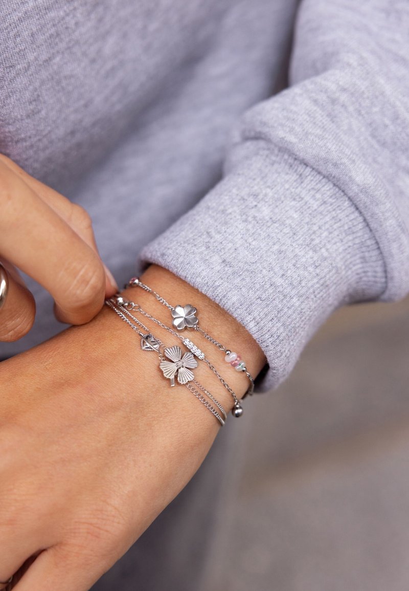 Bracciali metallici argento con accenti floreali, catene e perline in delicati colori pastello, indossati su un polso accanto a un maglione grigio.