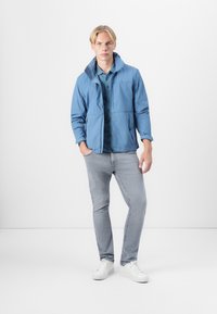 Lyseblå jakke med krage, utstyrt med glidelås og sidelommer, båret over en mørkeblå polo-shirt, sammen med lysegrå jeans og hvite joggesko.