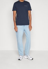 Marinblå bomullströja med kort ärm och en liten vit logotyp på bröstet, i kombination med ljusblå raka jeans och vita sneakers.