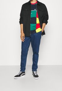 Camisa negra de cremallera sobre una camiseta a rayas multicolores, combinada con pantalones de mezclilla azules y zapatillas negras y blancas, mostrando un atuendo casual.