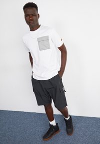 Hvid t-shirt med lys grå lomme, sorte cargoshorts og sorte høj-top sneakers med brune såler, sat mod en neutral baggrund.