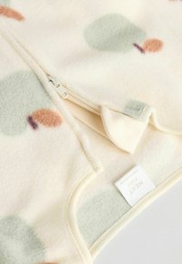 Ljus krämfärgad fleece tyg med dragkedja. Har dämpade gröna och persikofärgade trädformer. Mjuk textur och rundad krage. Varumärkesetikett synlig.