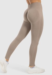 Beige high-waisted leggings met een getailleerd ontwerp; textiel met verticale lijnen en naadaccenten. Draagt met witte sportschoenen.