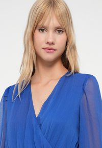 Tânără cu păr blond drept, purtând o bluză bleu transparentă cu decolteu în V, privind spre cameră pe un fundal alb simplu.