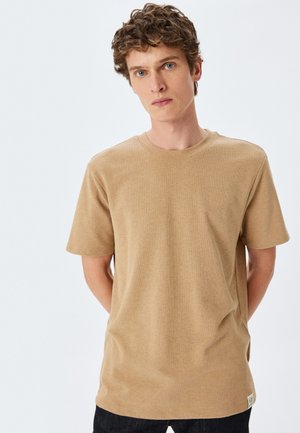 Beige T-Shirt mit kurzen Ärmeln aus strukturiertem Stoff und klassischem Rundhalsausschnitt, mit einem kleinen Etikett unten links.