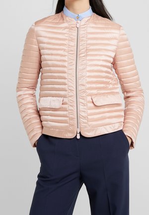 Femme portant une veste matelassée rose clair avec une fermeture éclair à l'avant, des poches à rabat, un pantalon bleu marine et une chemise bleu clair à col.