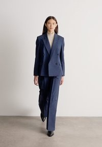 Žena kráčející vpřed, oblečená v tmavě modrém dvouřadém obleku s kalhotami širokých nohavic a světle šedém roláku, na pozadí bílé stěny.