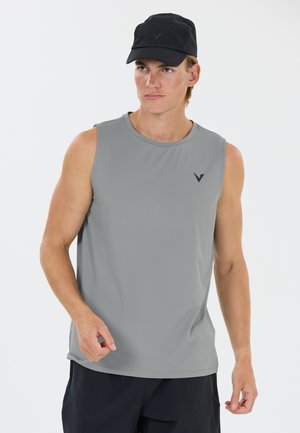Mann, der ein graues ärmelloses Shirt und eine schwarze Kappe trägt, steht vor einem schlichten weißen Hintergrund.
