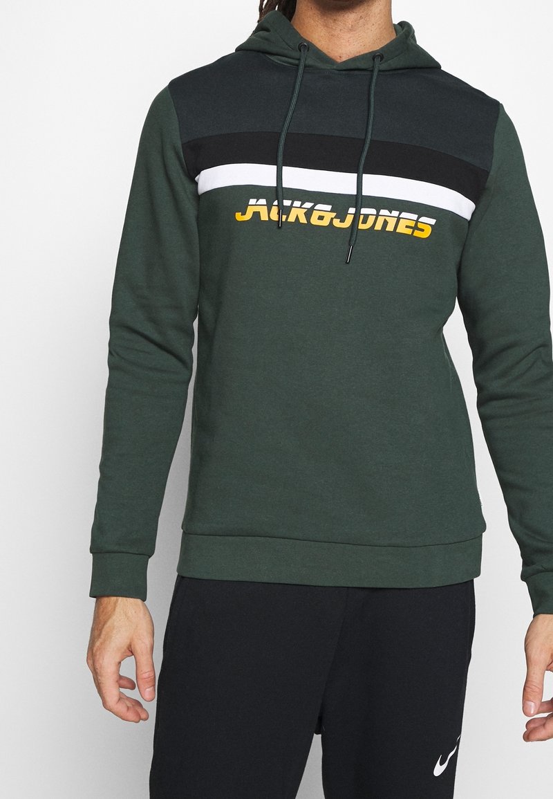 Zelená mikina s kapucí s designem v barevných blocích, obsahující černý, bílý a žlutý text "JACK&JONES." Vyrobena z jemné bavlněné směsi, s texturovaným povrchem.