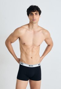 Schwarze Boxer-Brief mit einem weißen, gebrandeten Bündchen. Der Stoff wirkt glatt und körperbetont, geeignet für Freizeitkleidung. Das Modell steht mit den Händen in den Hüften.