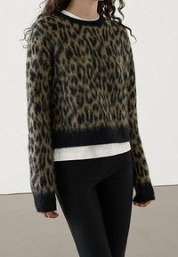 Leopardi-kuosiset neuleet pehmeästä, pörröisestä materiaalista mustilla yksityiskohdilla. Siinä on lyhyt malli ja pitkät hihat, kerroksittain valkoisen topin päällä.