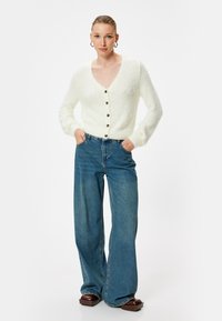 Cardigan bianco in peluche con scollatura a V e bottoni, abbinato a jeans in denim blu a gamba larga e scarpe con zeppa marroni.