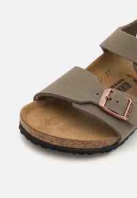 Sandal s smeškom od smešnog pluta s dva maslinasta remena. Uključuje metaliziranu bakrenu kopču. Ima teksturirani gornji dio i crnu gumenu potplatu.