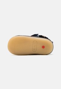 Sandale noire avec une semelle en caoutchouc flexible, dotée d'un motif texturé, d'une sangle ajustable et d'un point rouge bien visible à la base.