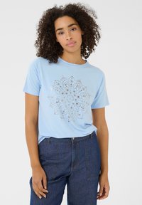 T-shirt bleu clair à manches courtes, col rond, orné d'un motif de mandala argenté au centre. Associé à un pantalon en denim foncé.