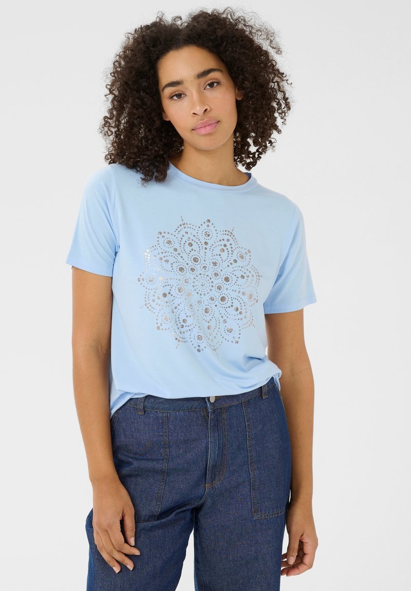 T-shirt bleu clair à manches courtes, col rond, orné d'un motif de mandala argenté au centre. Associé à un pantalon en denim foncé.