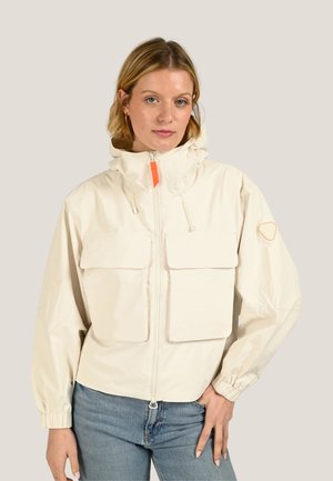 Femme portant une veste crème zippée avec de grandes poches avant et une capuche, associée à un jean bleu, debout devant un fond neutre.