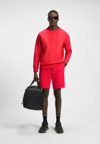 Maglione rosso con scollatura rotonda e piccolo logo, abbinato a pantaloni corti rossi coordinati. Zaino nero con forma curva e chiusura a zip.