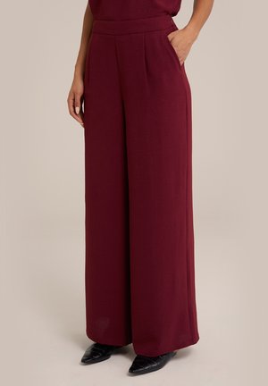 Frau trägt hoch taillierte, weit geschnittene burgunderrote Hosen mit Seitentaschen, kombiniert mit schwarzen spitzen Schuhen, stehend vor beigem Hintergrund.