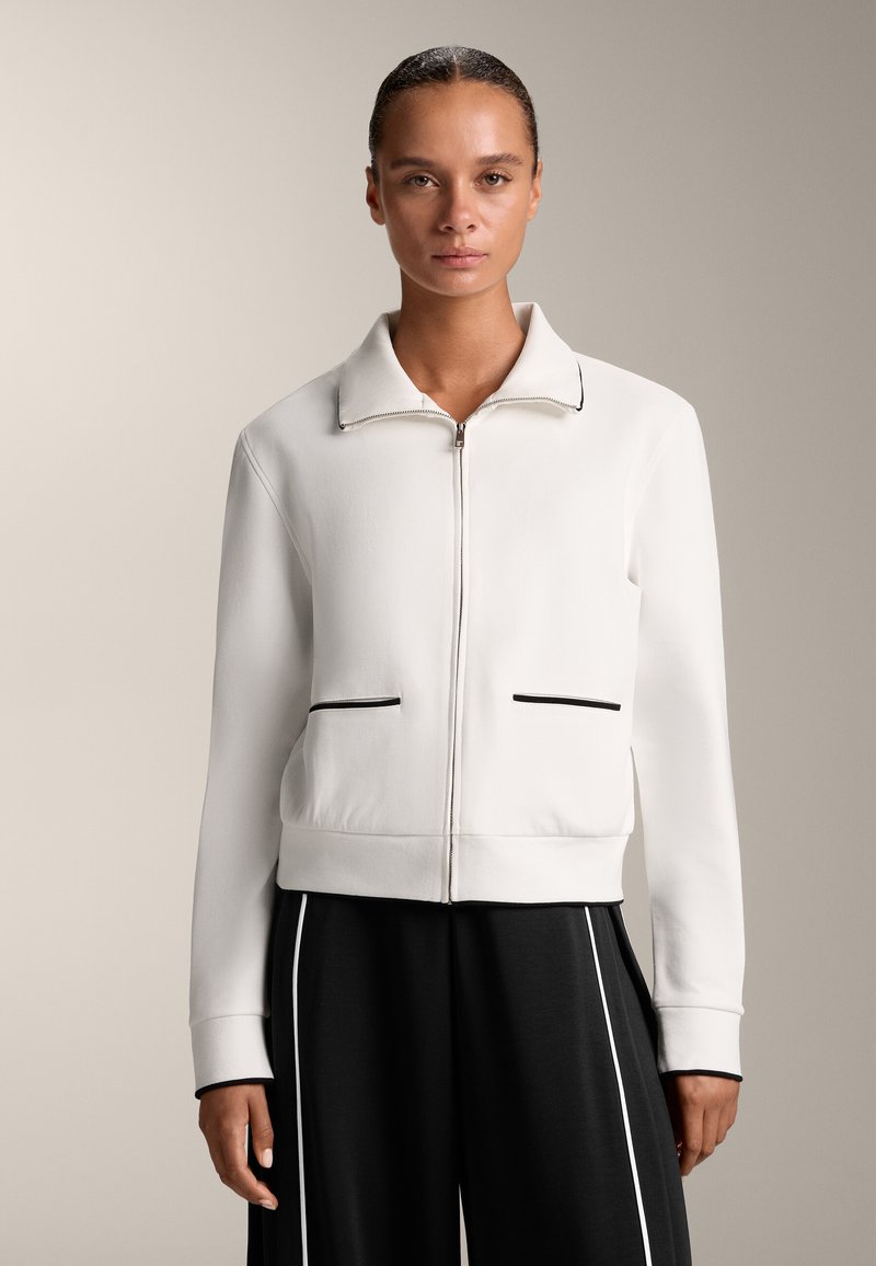 Fehér cipzáras dzseki lapos gallérral, két elülső zsebbel és fekete díszítésű részletekkel. Simító anyag, ami gondosan illeszkedik.